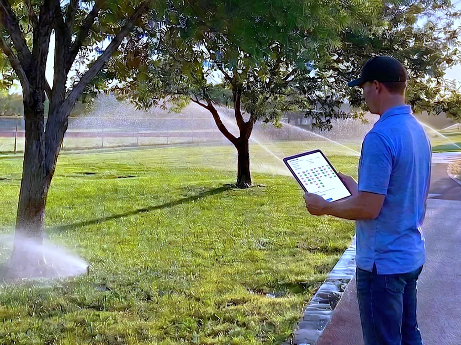 A technician logging findings on a tablet while a zone runs behind him, sprinklers throwing water across the lawn.